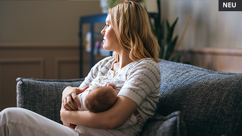 Mutter mit Neugeborenem auf dem Arm, sitzt im Wohnzimmer auf der Couch